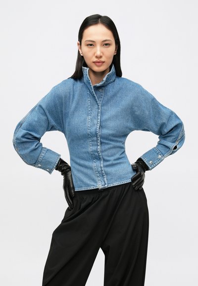 Denimjakk, millel on kõrge krae, nööbitav esiosa ja pikad varrukad. Hele sinine värv tikanddetailidega. Kandmiskõlblik mustade kinnastega.