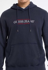Mees, kellel on seljas tumesinine kapuutsiga särk, millel on ees tasku ja millel on rinnal valgete ja punaste tähtede "GUESS JEANS" brodering.