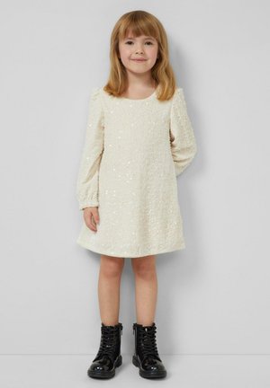 Robe couleur crème ornée de sequins, avec un col rond, des manches bouffantes et un ourlet évasé, associée à des bottines noires à lacets.