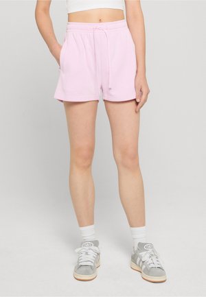 Femme portant un short à cordon de serrage rose clair, un haut court blanc, des chaussettes blanches montantes et des baskets Adidas grises, debout sur un fond uni.