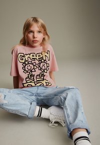 T-shirt rosa a maniche corte con la grafica nera del testo "Green" e due disegni di fiori. Indossato con jeans chiari strappati e sneaker bianche.