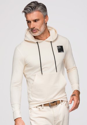 Homme aux cheveux gris et à la barbe portant un sweat à capuche crème avec des cordons noirs et un pantalon beige, regardant vers le bas à sa gauche.