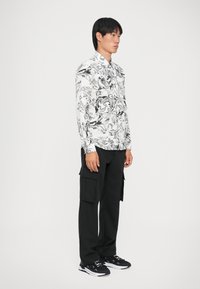 Camicia a fantasia in bianco e nero con disegni a vortice, abbinata a pantaloni cargo neri con più tasche e scarpe da ginnastica nere.