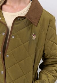 Olīvu zaļa izšūta jaka ar brūnu kordurojas apkakli, spiedpogām un nelielu rakstzīmes izšuvumu.