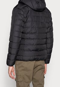 Doudoune noire avec sections matelassées et capuche, dotée d'une finition brillante et de manches élastiques, portée avec un pantalon cargo kaki.