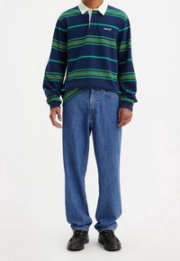 Námořnické pruhované polo tričko s dlouhým rukávem v zelenožlutých pruzích, bíločervený límec, v kombinaci s volnými modrými džínami a černými teniskami.
