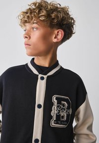 Chaqueta varsity en negro y beige con cuello redondo, cierre de botones y detalle de logotipo bordado en el pecho. Textura de tela suave.