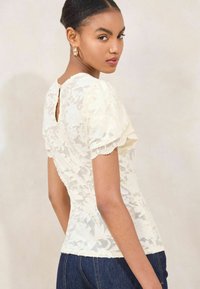 Lace top in off-white with short puff sleeves, scalloped edges, and a keyhole back closure. Worn with dark denim.
