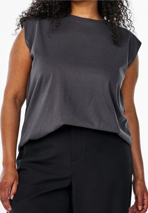 Woman wearing dark gray sleeveless top and black pants, curly hair visible, shown from shoulders to hips against white background.