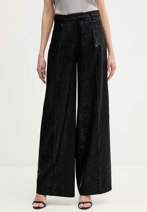 Donna che indossa pantaloni neri a vita alta e gamba larga con una texture, abbinati a sandali neri con punta aperta su uno sfondo semplice.