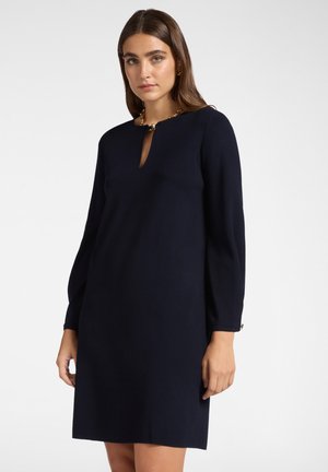 Donna che indossa un semplice vestito nero a maniche lunghe con scollatura a chiave e dettagli in catena dorata su uno sfondo bianco.