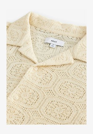 Kermainen neulepolo, jossa on kuvioitu pinta, kaulakotelo, nappisuljenta ja merkkilogo sisäpuolella.