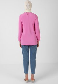Maglietta rosa a maniche lunghe con dettaglio a chiave sulla schiena, abbinata a jeans azzurri slim-fit e sandali beige, su uno sfondo neutro.