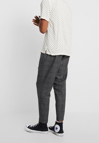 Man med grå rutiga tapered byxor, vit mönstrad skjorta med kort ärm och svarta högskaftade Converse sneakers, står med händerna ihopfoldade.