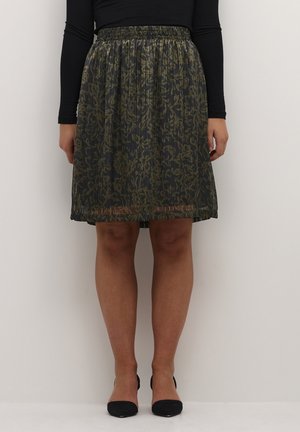 Frau trägt einen knieumspielenden, dunklen, geblümten Faltenrock mit elastischem Bund, kombiniert mit einem schwarzen Langarmoberteil und schwarzen High Heels.