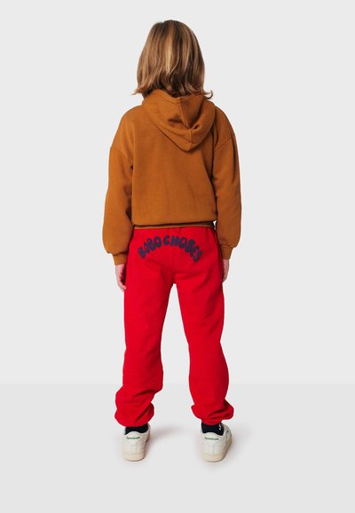 Brauner, kurzer Kapuzenpulli mit Kapuze, kombiniert mit leuchtend roten Trainingshosen, die auf der Rückseite navyblauen Schriftzug tragen. Legere Schuhe runden das Outfit ab.
