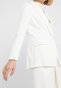 Blazer bianco su misura con una leggera lucentezza, spalle strutturate, chiusura con un solo bottone e rever eleganti. Abbinato a una camicetta color crema.