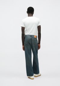 Jeans bleu foncé à coupe droite, avec poches arrière et un patch de marque en cuir marron clair. Portés avec un t-shirt blanc et des baskets blanches.