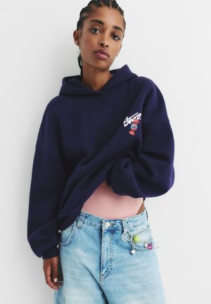 Hoodie surdimensionné bleu marine avec une poche avant, présentant un design graphique coloré. Associé à un short en jean bleu clair et des accessoires.