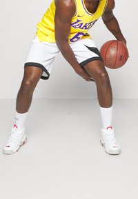 Gul baskettröja med lila accenter, svartvita shorts och vita sneakers med röda detaljer. Spelare som dribblar en basketboll.