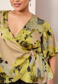 Blumenwrap-Bluse in Gelb- und Grüntönen mit kurzen, schwingenden Ärmeln und einem Bindeband an der Taille; aus einem glatten, leichten Stoff gefertigt.