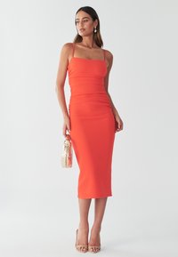 Orangenes Midi-Kleid mit Spaghetti-Trägern, Rüschendetail und figurbetontem Schnitt. Kombiniert mit einer metallisch goldenen Handtasche und nude-farbenen High Heels.