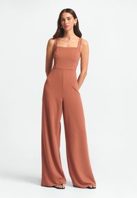 Terrakotta jumpsuit med fyrkantig halsringning, tjocka axelremmar, vida ben och slät tygkvalitet med minimala sömmar.