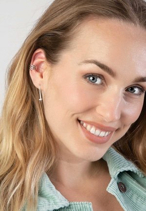 Zilveren oorbellen met een delicate ketting en een klein kruisjeshangertje. Het haar is losjes gestyled en de persoon draagt een lichtgroene corduroyjas.