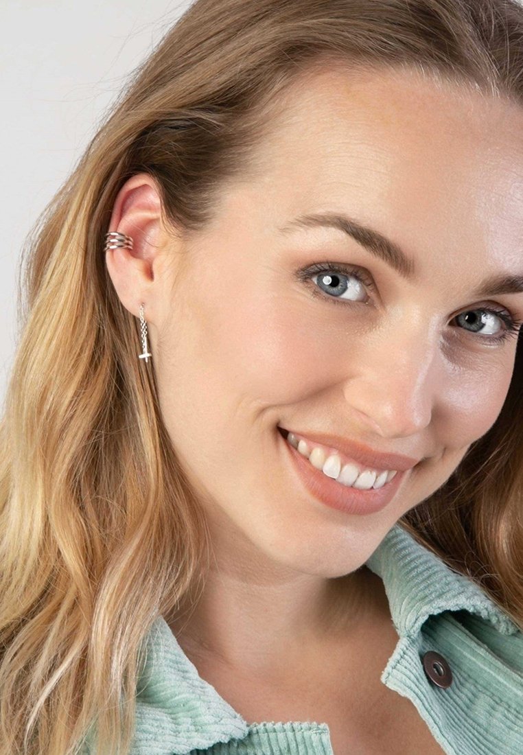 Zilveren oorbellen met een delicate ketting en een klein kruisjeshangertje. Het haar is losjes gestyled en de persoon draagt een lichtgroene corduroyjas.