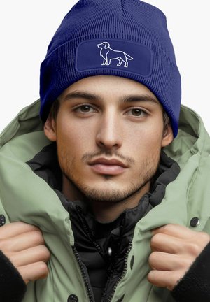 Blue knit beanie with a textured finish and white dog silhouette patch, paired with a green puffer jacket, enhancing warmth and style.
