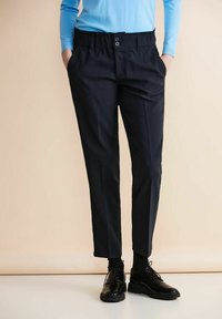 Skräddarsydda marinblå byxor med en mjuk textur, med en elastisk midja, två fickor och två knappar fram. Bärs med svarta skor.