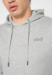 Grijze capuchontrui van zachte stof. Heeft een logo op de borst, een verstelbare trekkoord en metalen uiteinden op het trekkoord.
