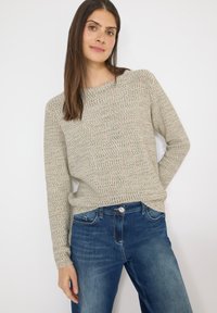Femme aux longs cheveux bruns portant un pull en maille gris clair et un jean bleu, debout devant un fond blanc.