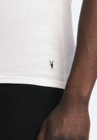 Hvid ribbet tanktop med en sort logoaccent nær kanten, båret over sorte bukser. Glat tekstur og tætsiddende design.