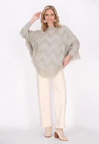 Poncho grigio chiaro con frange e motivo a zigzag, abbinato a pantaloni crema e stivali con tacco beige. Tessuto morbido e vestibilità rilassata.