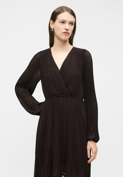 Γυναίκα που φορεί μακρυμάνικο μαύρο πλισέ φόρεμα με λαιμόκοψη σε σχήμα V, στέκεται με το ένα χέρι στη μέση της μπροστά σε απλό φωτεινό φόντο.