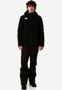 Giacca nera impermeabile con cappuccio, zip frontale e logo bianco. Abbinata a pantaloni neri strutturati con tasche con zip.