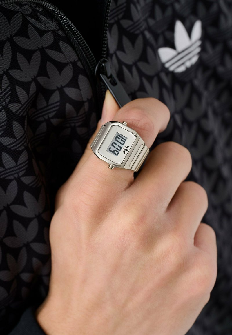 Main tenant la fermeture éclair d’une veste noire Adidas, portant une montre digitale argentée en forme d’anneau affichant 6h02 et le logo Adidas à l’écran.