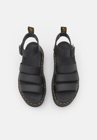 Sandales en cuir noir avec trois larges brides, boucle de cheville ajustable et coutures jaunes contrastantes le long de la semelle.