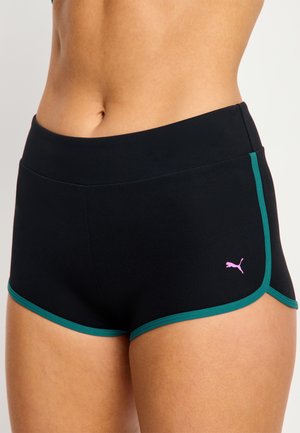 Gros plan d'une personne portant un short de sport noir avec des bordures vertes et un petit logo Puma rose sur le côté gauche.