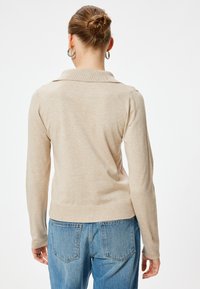 Beżowa bluzka z długim rękawem i kołnierzykiem, o gładkiej fakturze i prążkowanym wykończeniu. Połączona z niebieskimi dżinsami z wysokim stanem.