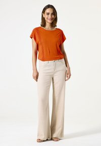 Top corto de manga corta naranja con costuras texturizadas, combinado con pantalones de pierna ancha en un tono beige claro. Diseño minimalista, tejido suave y zapatos planos.