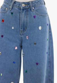 Vaqueros de mezclilla azul decorados con adornos de gemas multicolores de diversas formas dispersas por la parte frontal de las piernas.