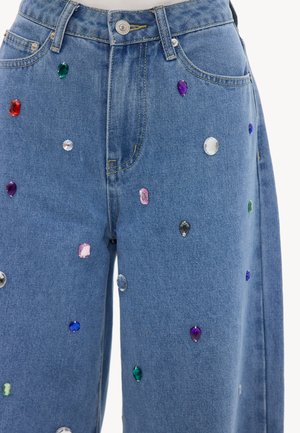 Jean en denim bleu orné de pierres précieuses multicolores de formes variées dispersées sur le devant des jambes.