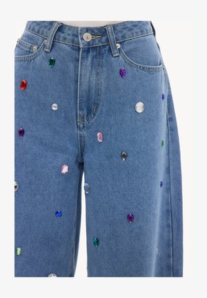 Jean en denim bleu orné de pierres précieuses multicolores de formes variées dispersées sur le devant des jambes.