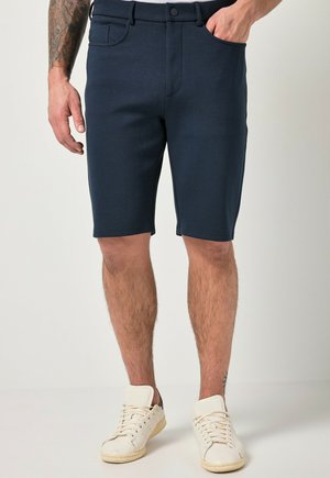 Mand iført mørkeblå shorts med lommer og hvide sneakers, stående med den ene hånd i lommen mod en ensfarvet lys baggrund.