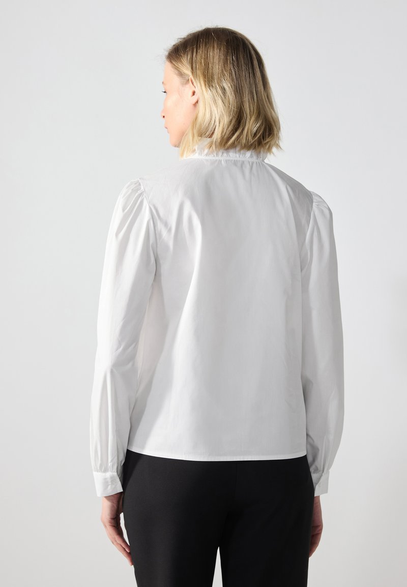 Chemise blanche à boutons avec des manches bouffantes, texture lisse et col arrondi. Portée avec un pantalon noir, mettant en valeur un design simple et épuré.