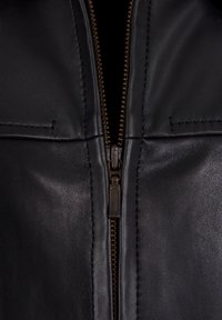 Zwarte leren jas met een bronzen rits, met een soepele textuur, dubbele borstzakken en een strakke, minimale stiksel-detail.