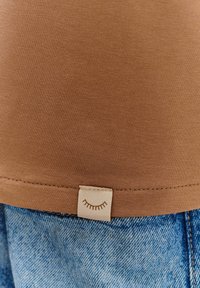 Hnedý rebrovaný tielko s prešitým lemom, s malou béžovou nálepkou na spodku s jednoduchou grafikou oka, skombinované s modrými džínsami.