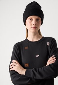 Felpa nera con vari toppe ricamate in tonalità di marrone e rosa. Un berretto di maglia nero indossato sulla testa completa il look.